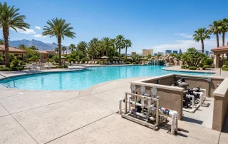 A bright, photorealistic wide shot of a large commercial resort swimming pool in the Las Vegas Valley at midday. The water is crystal clear and calm. The surrounding deck and concrete are fully intact, with no trench, no dirt pile, and no digging equipment in sight. Palm trees, a desert mountain skyline, and a clear blue Nevada sky in the background. A small, tidy pipe injection rig with pressure gauges rests on the pool deck near the equipment pad. Warm natural light, sharp focus, no people in frame. Shot on a DSLR, 24mm lens, clean professional tone. -